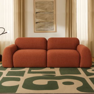 Two-section orange sofa in a neutral living room with a geometric green rug and beige curtains on either side.