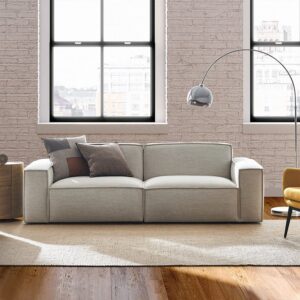 Light gray sofa with three throw pillows in a sunlit loft-style living room with exposed brick walls and large black-framed windows