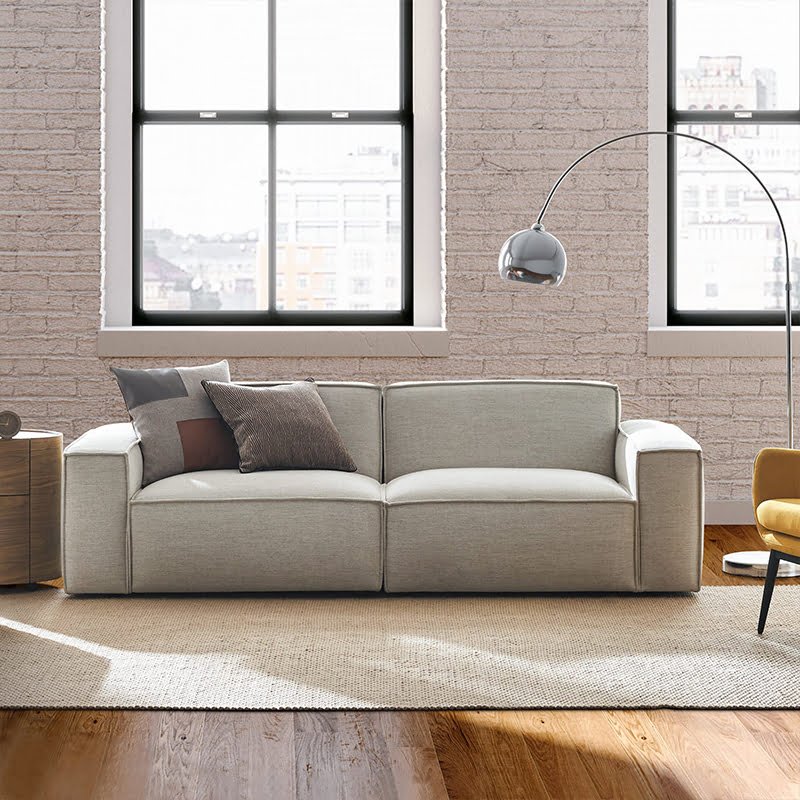 Light gray sofa with three throw pillows in a sunlit loft-style living room with exposed brick walls and large black-framed windows